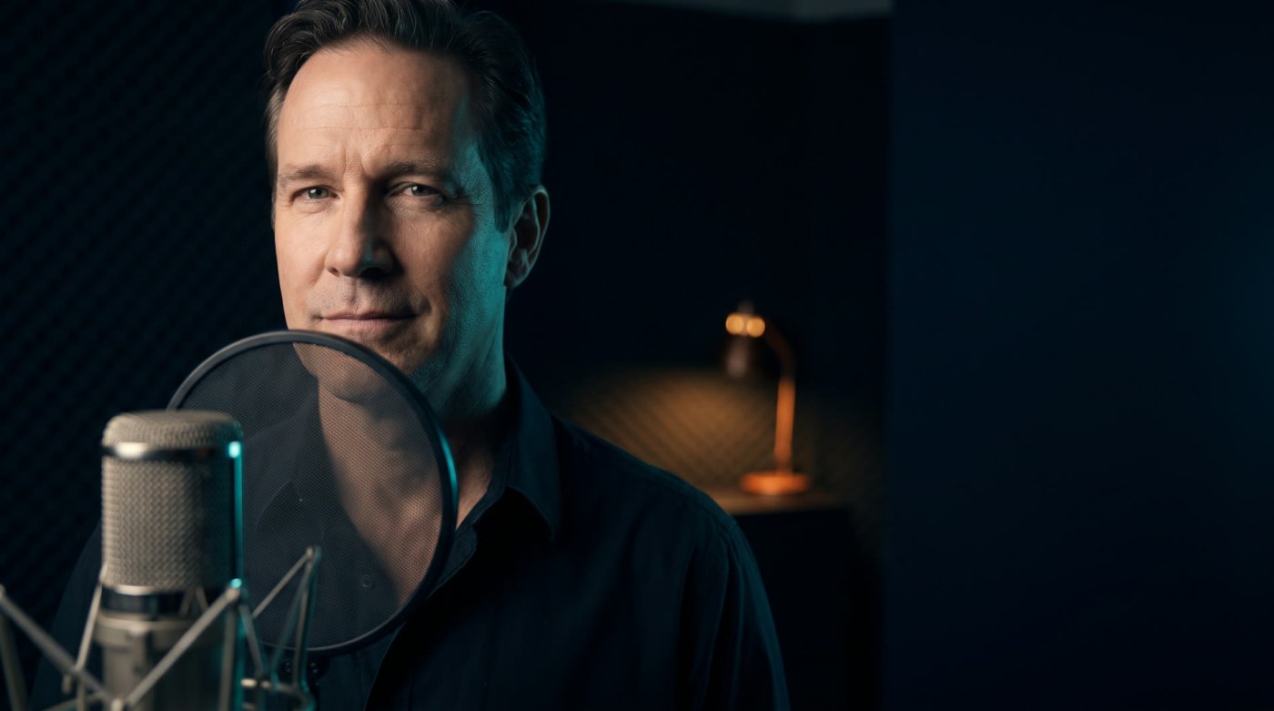 A voice actor at a large diaphragm studio microphone with a pop filter, in a dark booth lit by a warm desk lamp